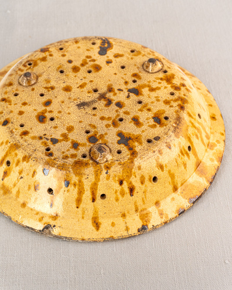 Antique French glazed earthenware fruit strainer with rust spots, small holes, and four base rivets on a light gray surface.