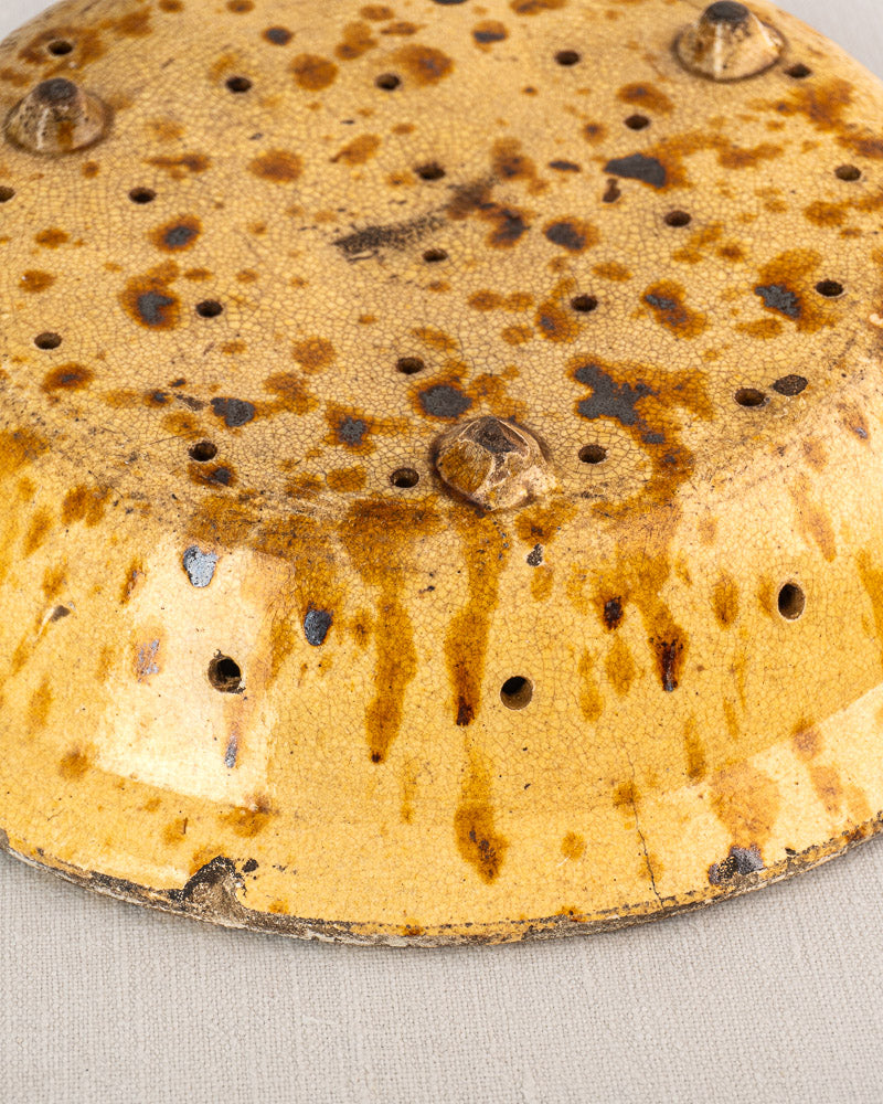 Upside-down 19th-century French glazed earthenware fruit strainer with brown spots on a light fabric background.