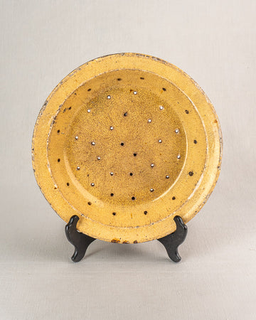 Antique French glazed earthenware égouttoir, round yellow plate with patterned holes, upright on a black stand.