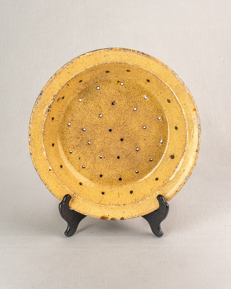 Antique French glazed earthenware égouttoir, round yellow plate with patterned holes, upright on a black stand.