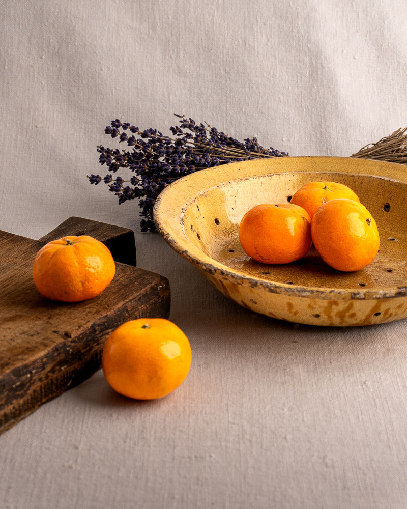 19th-century French glazed earthenware fruit strainer with tangerines, on fabric near dried lavender and a wooden board.