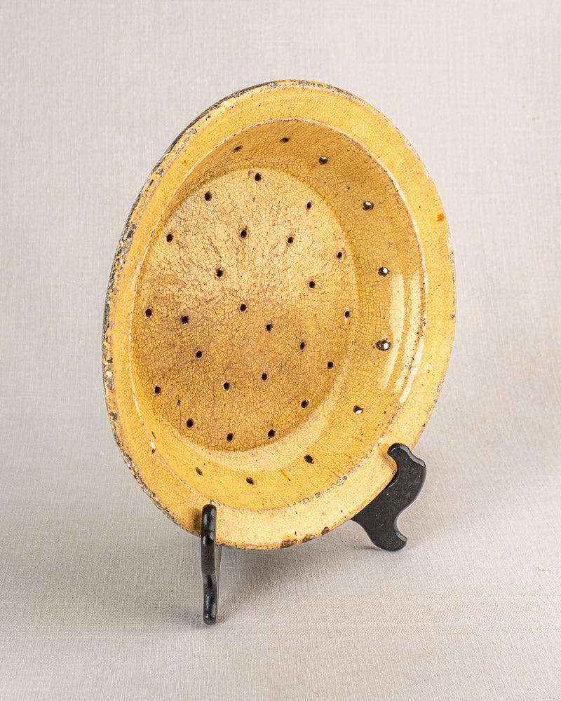19th-century French yellow glazed earthenware fruit strainer with small holes on a black metal stand against a neutral backdrop.