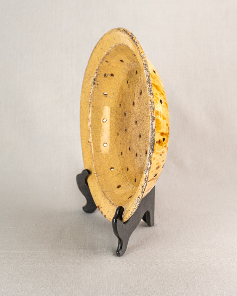 19th-century French glazed earthenware fruit strainer with small holes, speckled yellow glaze, on black stand.