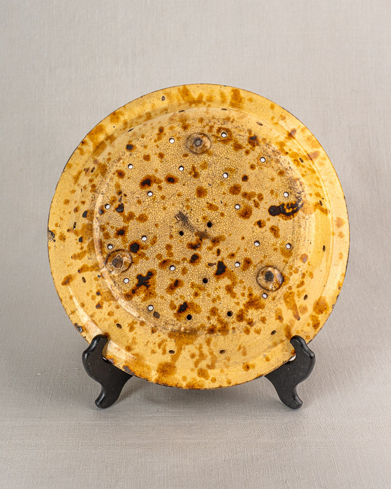Antique French glazed earthenware fruit strainer with speckled yellow-brown finish, patterned drainage holes, on black stand.