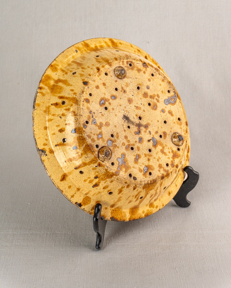 19th-century French glazed earthenware fruit strainer with yellowish-brown speckled finish, on black stand, gray background.