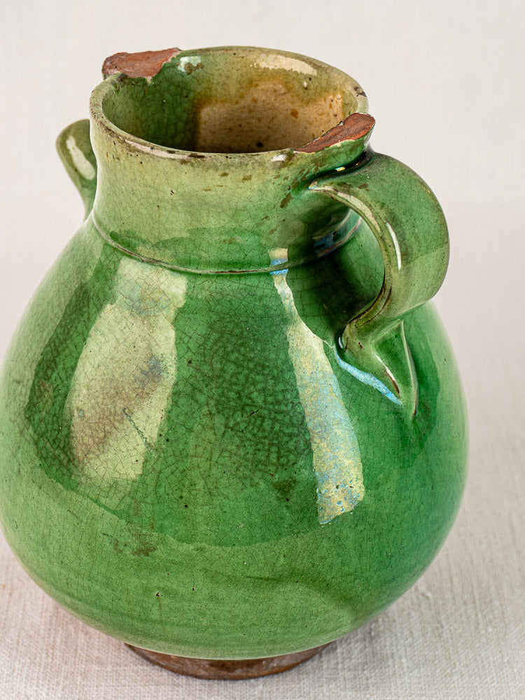 Antique French green glazed terracotta gargoulette with large handle, chipped rim, on neutral fabric.