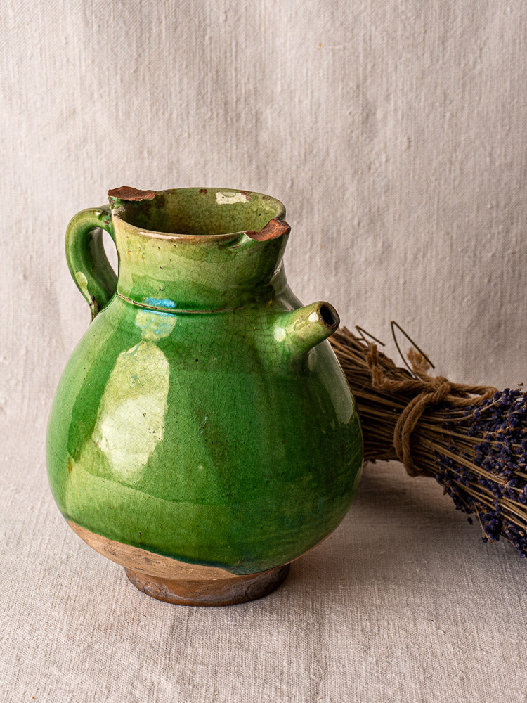 Antique French gargoulette water jug in green-glazed terracotta, chipped rim, on neutral fabric.