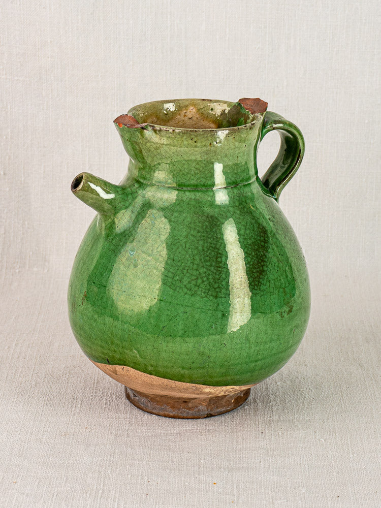 Antique French gargoulette water jug in green-glazed terracotta, chipped rim, on neutral fabric.