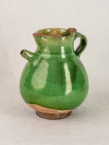 Antique French gargoulette water jug in green-glazed terracotta, chipped rim, on neutral fabric.