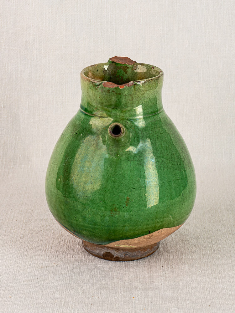 Antique French green-glazed terracotta gargoulette with chipped rim on neutral fabric background.