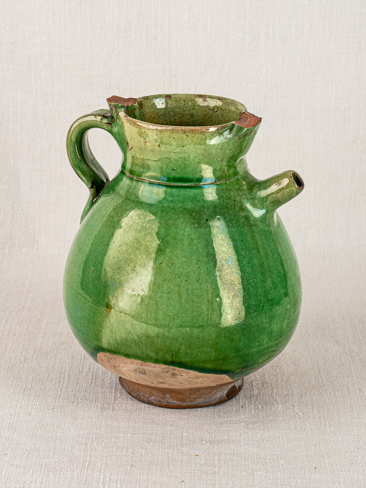 Antique French green-glazed terracotta gargoulette with single handle, spout, and chipped rim on fabric background.