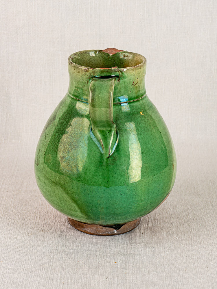 Antique French green glazed terracotta gargoulette with wide body and handle on neutral fabric background.