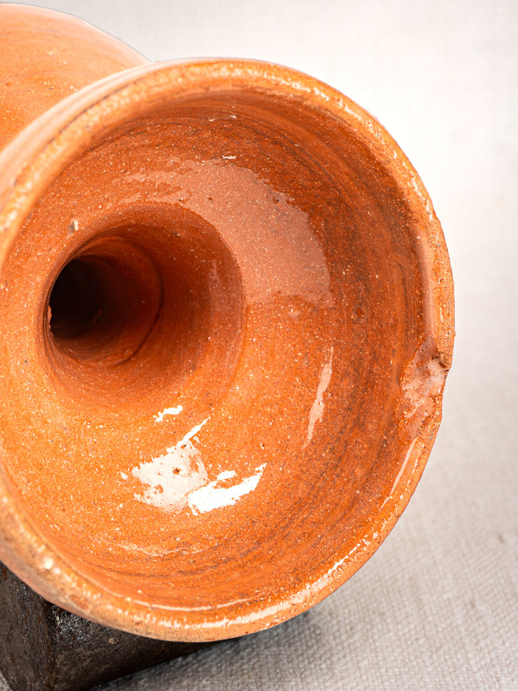 Antique French mammiforme terracotta jug with glossy orange-brown glaze and chipped rim, on light fabric.