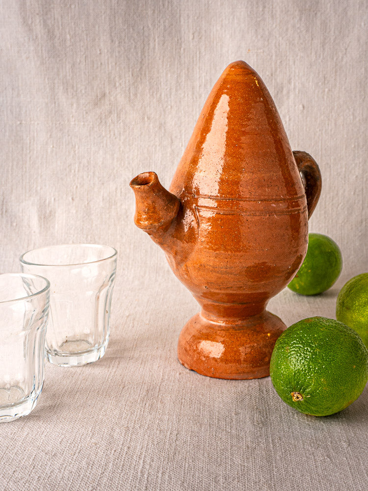 Antique French brown terracotta jug with spout beside two glass cups and three limes on fabric.