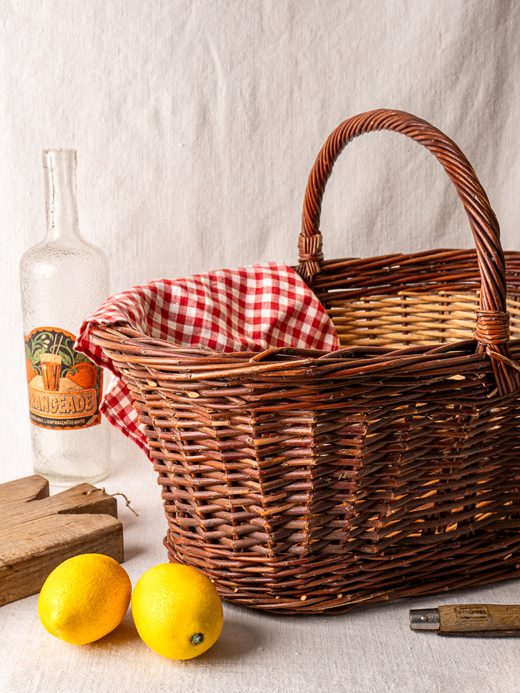 Antique French two-tone willow gathering basket with red and white checked cloth, beside lemons and clear bottle on neutral fabric.