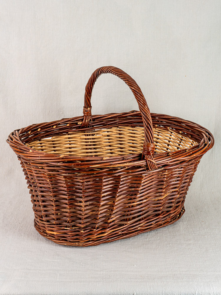 Antique French gathering basket circa 1900 in two-tone willow with handle, displayed on light fabric surface.