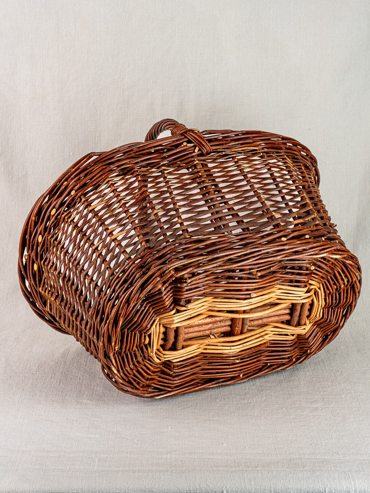 Antique French willow gathering basket, two-tone oval design with handle, on light gray fabric background.