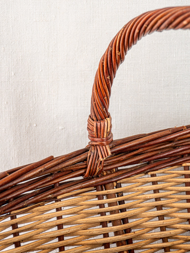 Antique French gathering basket with two-tone willow handle and intricate weave, set on a neutral fabric background.