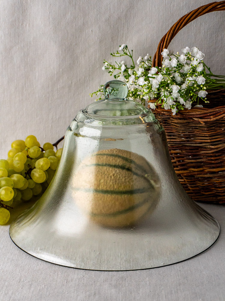 Antique French hand blown glass cloche covering a melon beside green grapes and basket of white flowers.