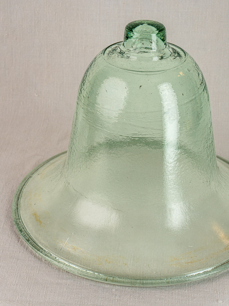 Hand-blown 19th-century French glass melon cloche with green tint and round handle on neutral fabric background.