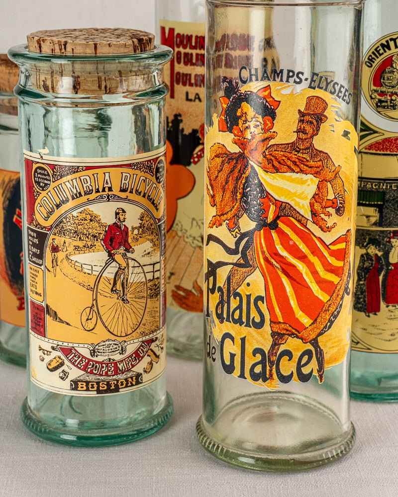 Set of five 1970s French glass jars with cork stoppers and Belle Époque poster labels grouped on a neutral surface.