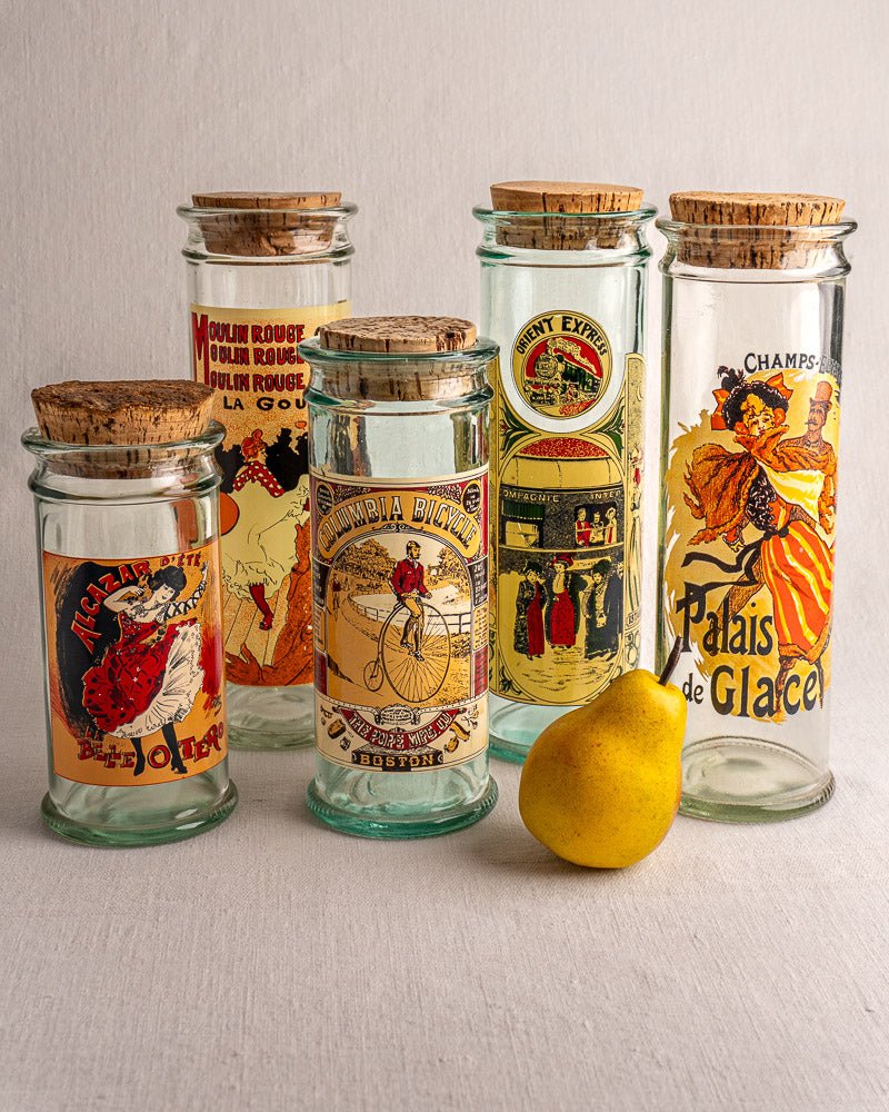 Set of five vintage French glass jars with cork stoppers beside a yellow pear on light surface.
