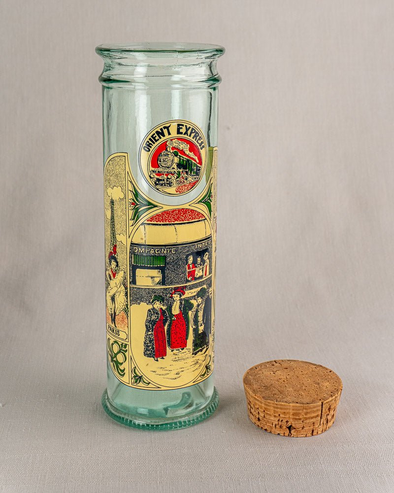 Tall clear glass jar with vivid Belle Époque Orient Express poster illustrations, cork stopper beside.