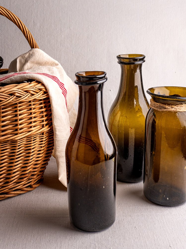 Three antique French brown glass bottles beside a wicker basket with white and red cloth.