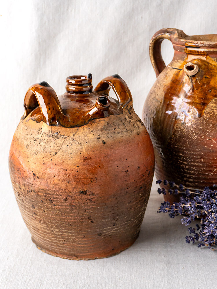 Antique French glazed terracotta oil jar with two handles and rustic brown finish, paired with a smaller three-spouted vessel on light fabric, dried lavender nearby.
