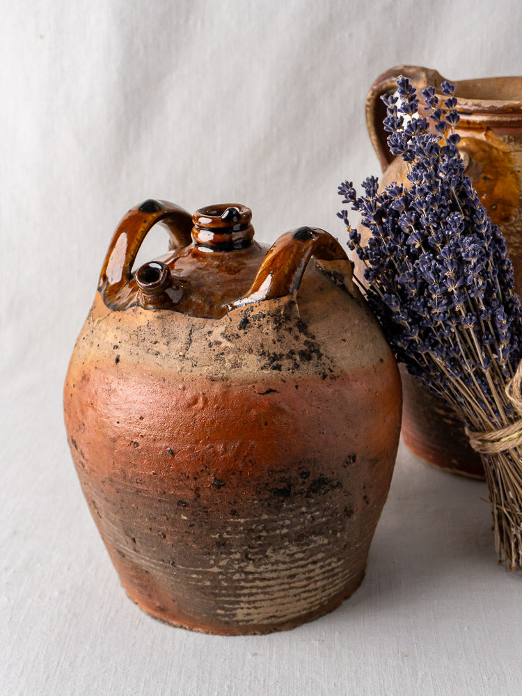 Antique French two-handled glazed terracotta oil jar beside dried lavender on neutral fabric.