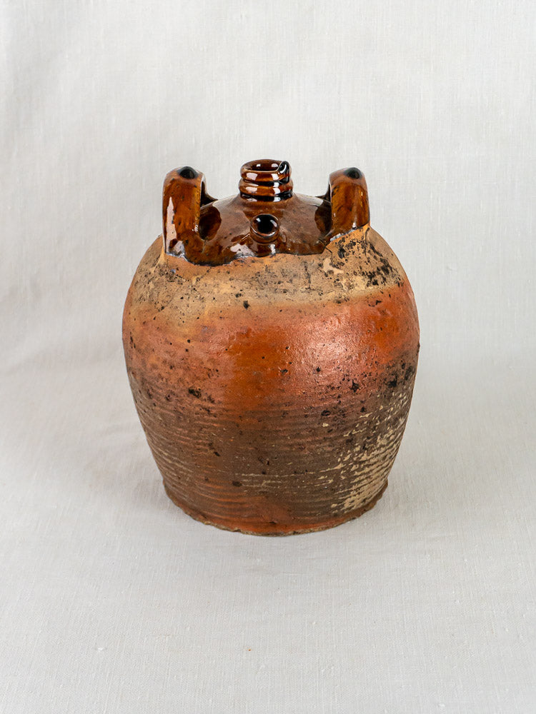 Antique French glazed terracotta oil jar with two handles and round body on light fabric.