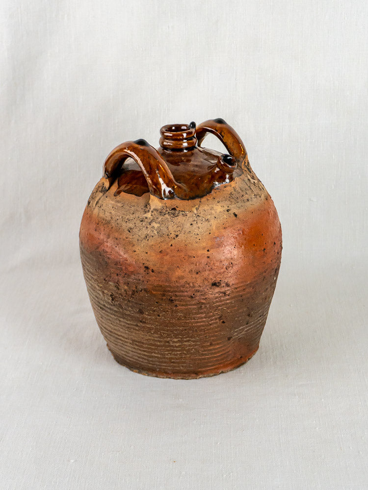 Antique French glazed terracotta oil jar with twin handles and small spout on light fabric.