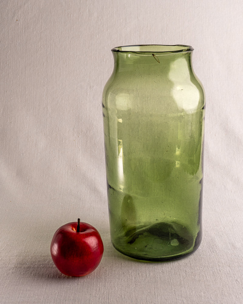 Antique French hand-blown green glass cornichon jar, late 18th century, beside a red apple on light fabric.