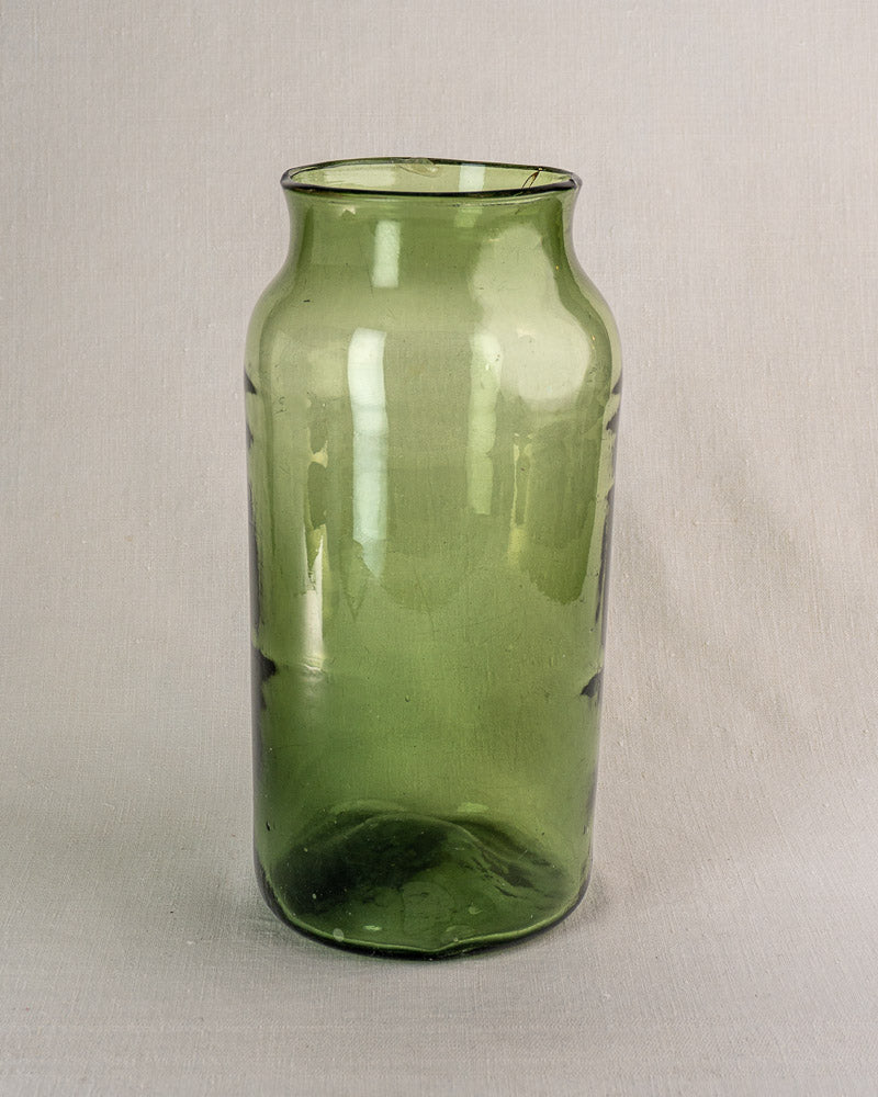 Antique French hand-blown green glass cornichon jar with flared rim on a light fabric background.