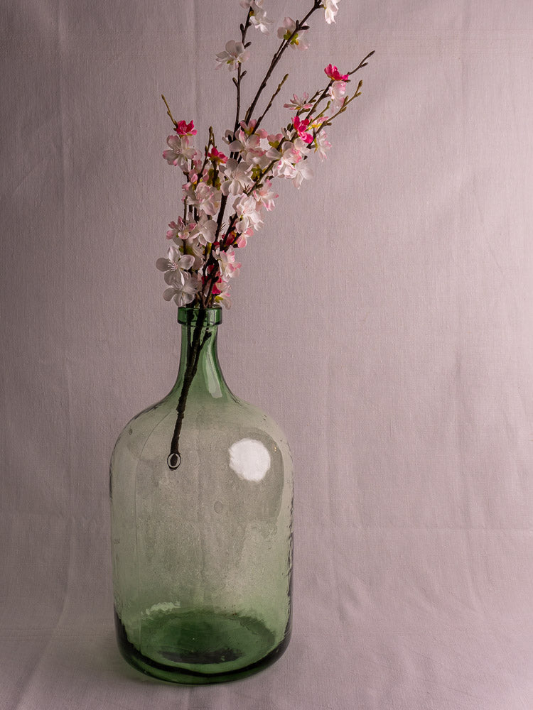Antique French green glass demijohn with pink and white blossoms, set against a plain light background.