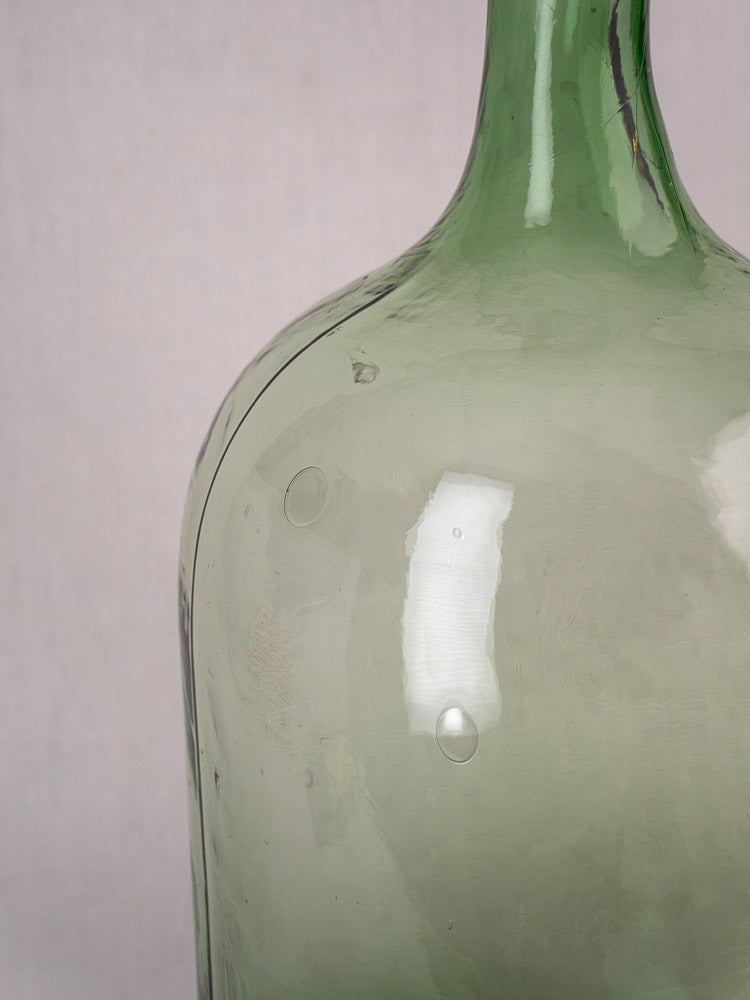 Antique French demijohn in hand-blown green glass with air bubbles, light reflections, neutral background.