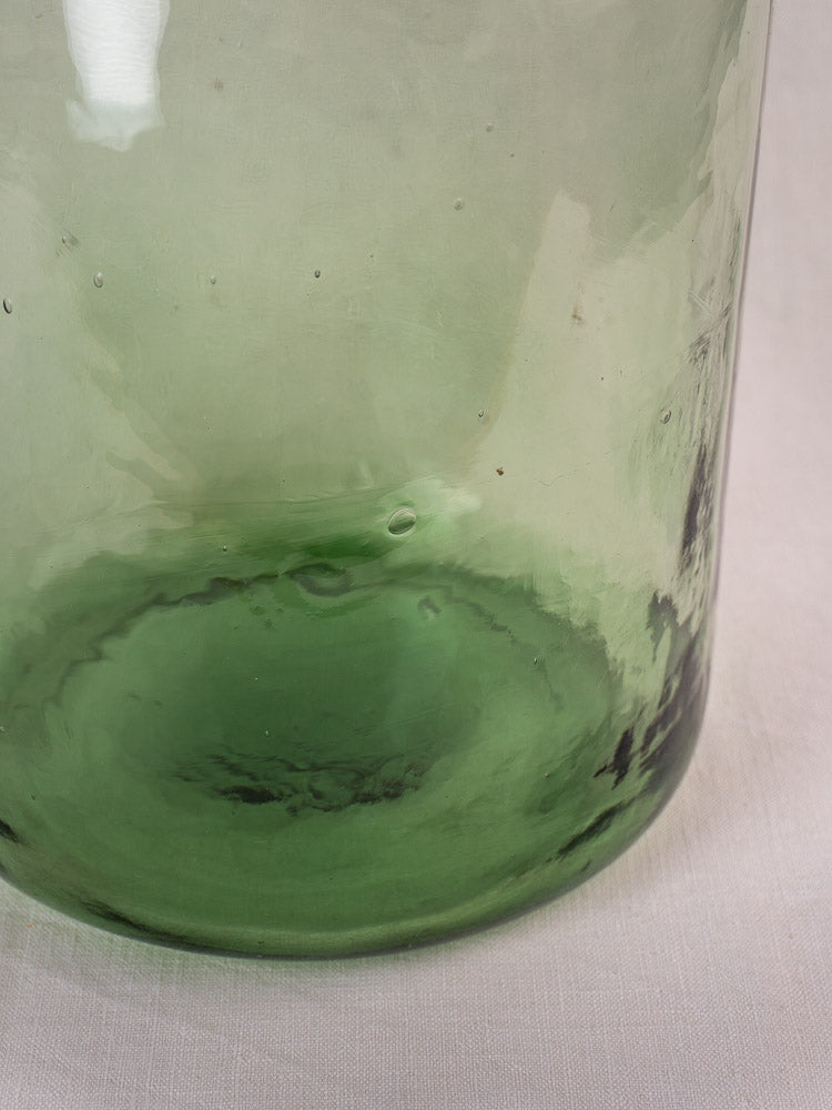 Antique French demijohn in hand-blown green glass, textured base with unique imperfections on a light fabric surface.