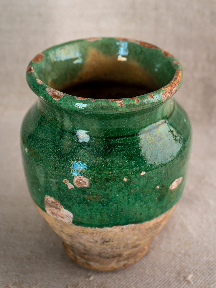 19th-century French confit pot with wide mouth, patchy green glaze, and weathered brown pottery on beige surface.