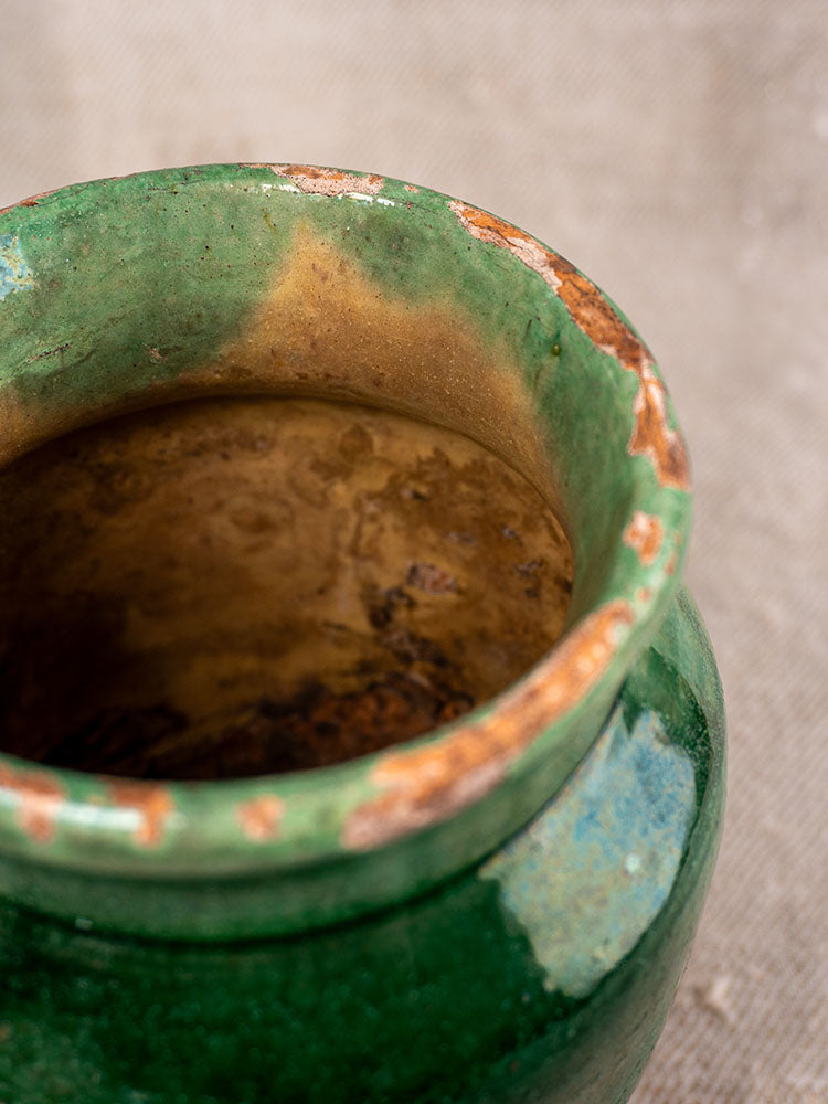 Antique French confit pot with green glaze, terracotta chips, and patina, set on a textured beige surface.