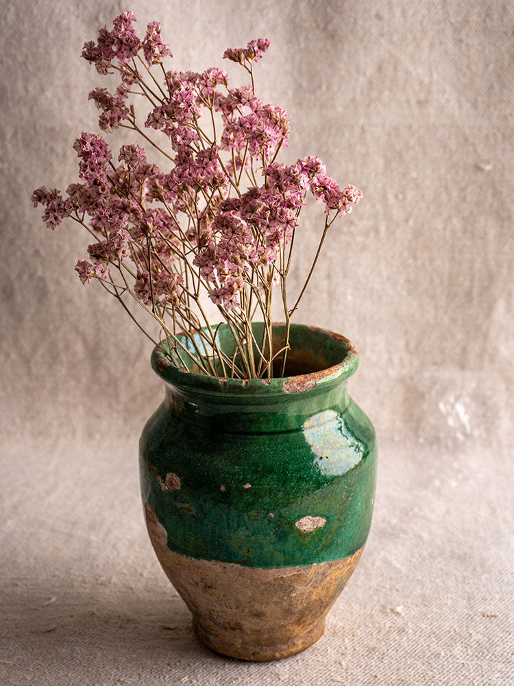 Antique French green glazed confit pot with dried pink flowers, set against textured beige fabric.