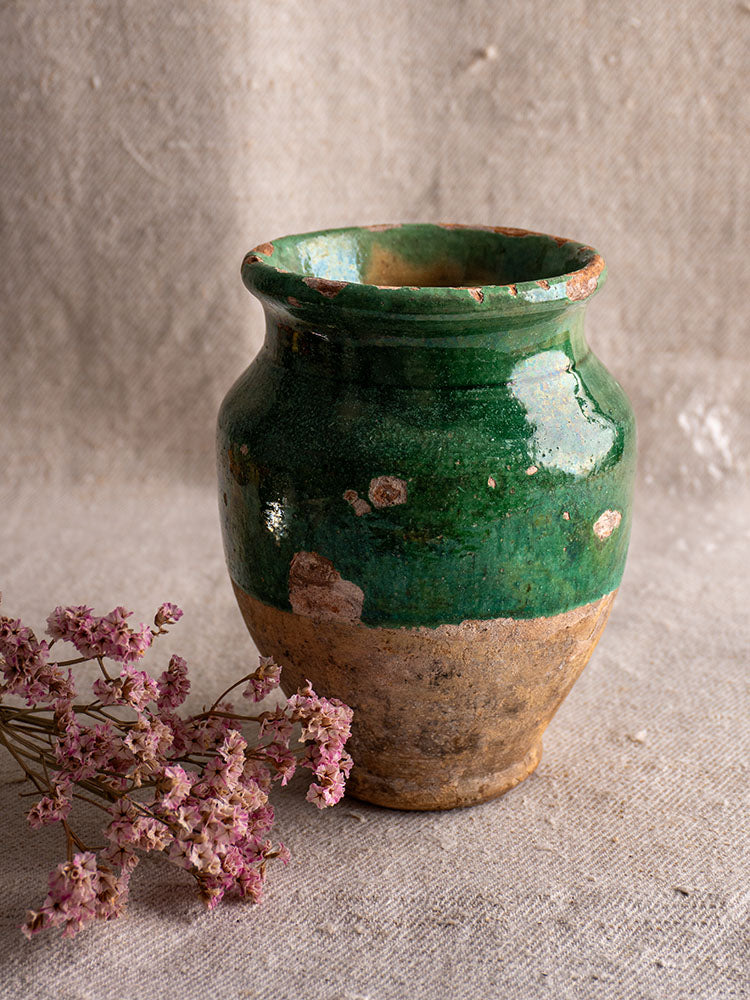 Antique French green-glazed confit pot with rustic ceramic surface beside dried pink flowers on textured fabric.