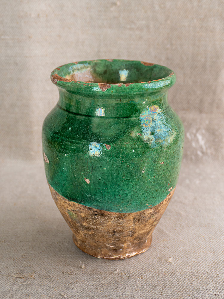 Antique French confit pot with rustic green glaze, chipped to reveal brown clay; wide mouth, rounded body on beige surface.