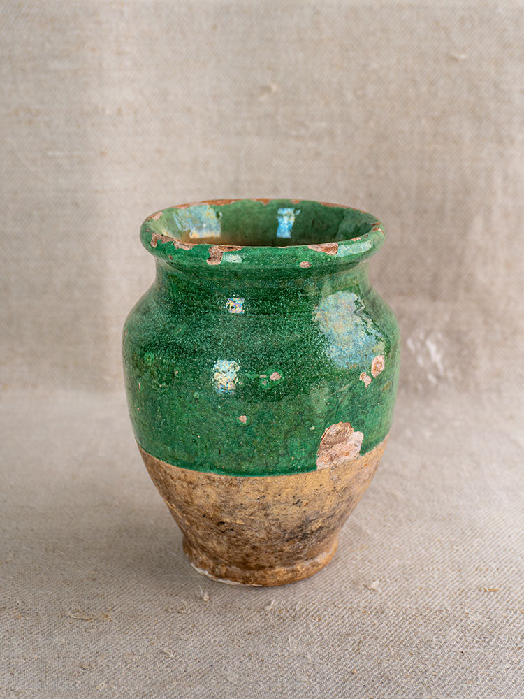 Antique French confit pot with chipped green glaze and unglazed brown terracotta, on textured beige fabric.