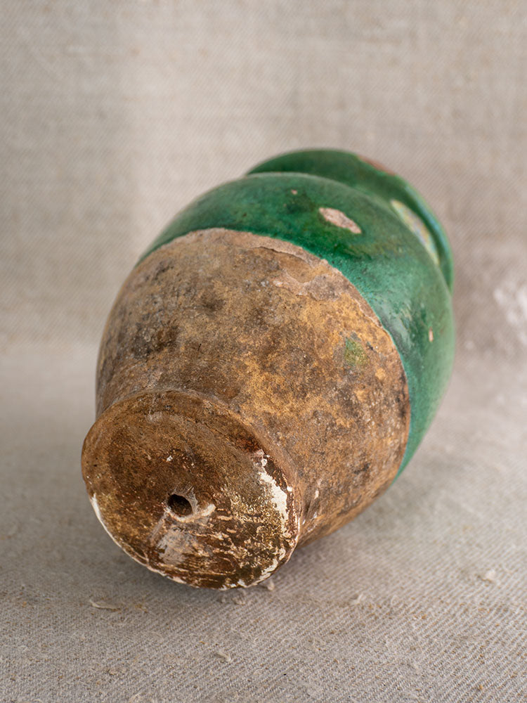 Antique French confit pot with green glaze, 19th century, showing wear and a small base hole on beige textured surface.