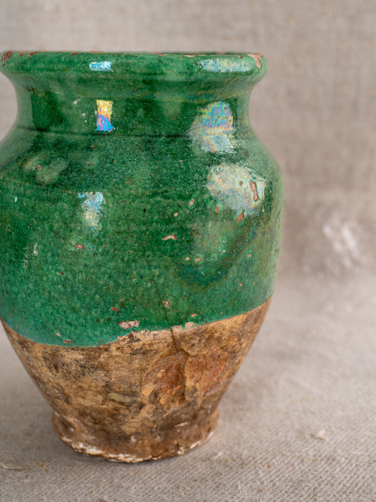 Antique French confit pot with glossy green glaze and brown base, showcased on a textured neutral background.