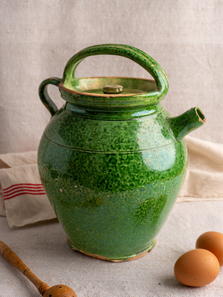 Antique French green glazed cruche orjol with lid and curved handle on beige fabric, eggs and honey dipper nearby.