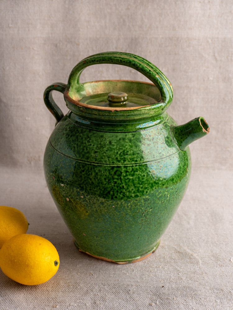 Antique French green glazed cruche orjol with curved handle and spout, displayed on beige textured surface with two lemons.