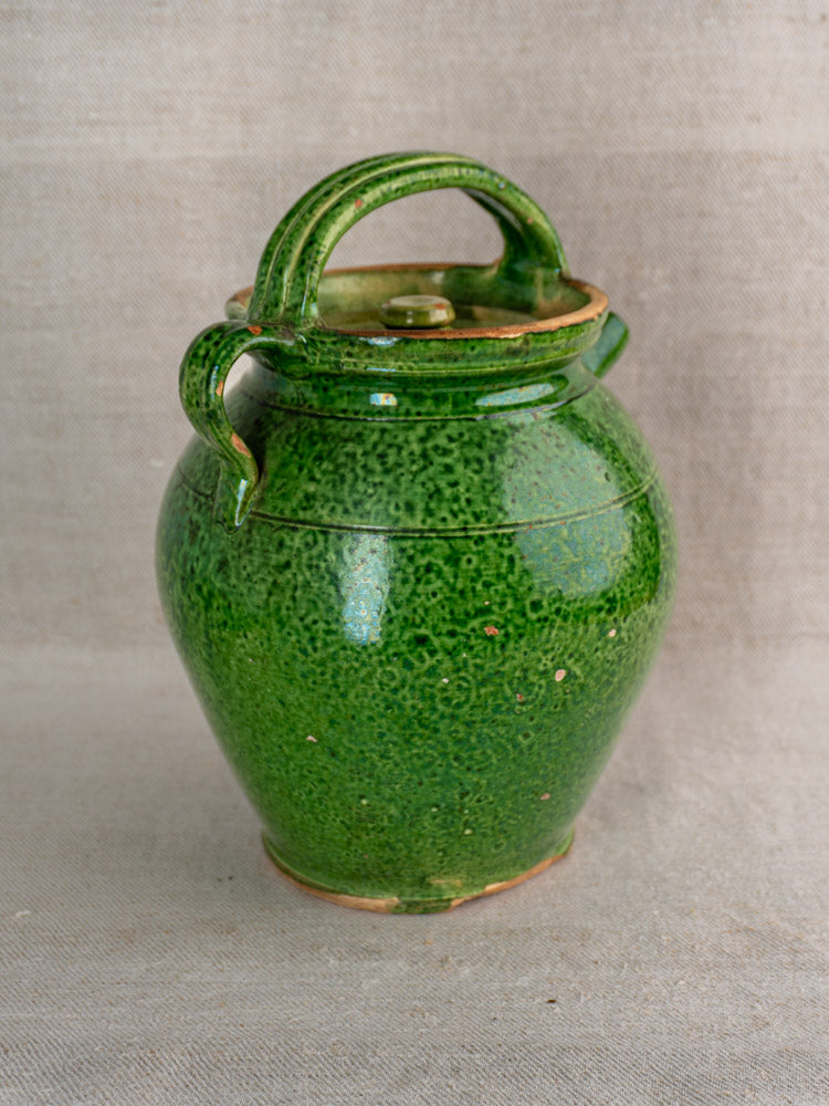 Antique French green glazed Cruche Orjol with two handles and lid, early 20th-century, on beige fabric.