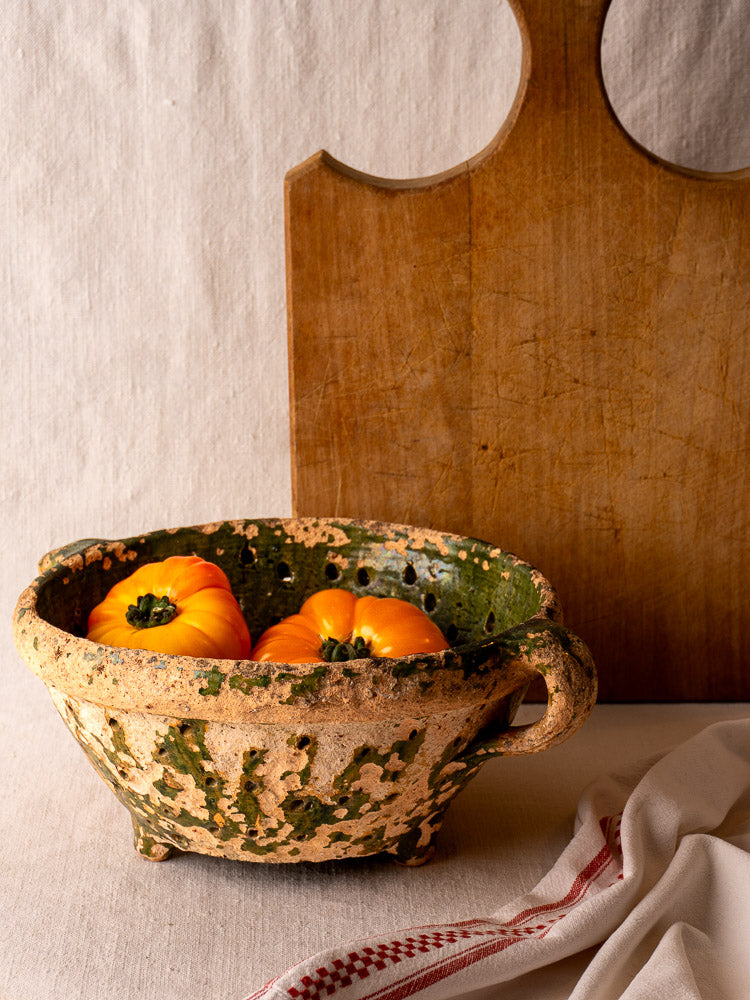 Late 18th-century French green glazed terracotta colander with three orange heirloom tomatoes, wood board and striped towel nearby.