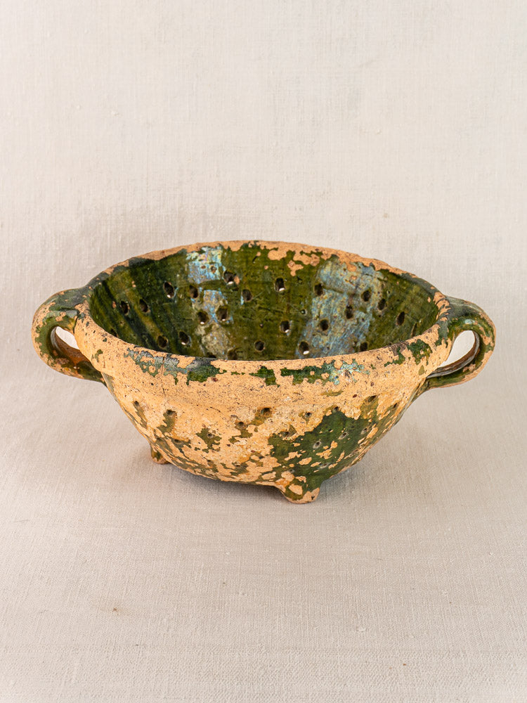 Antique French green glazed terracotta colander with two handles, speckled finish, on neutral fabric background.